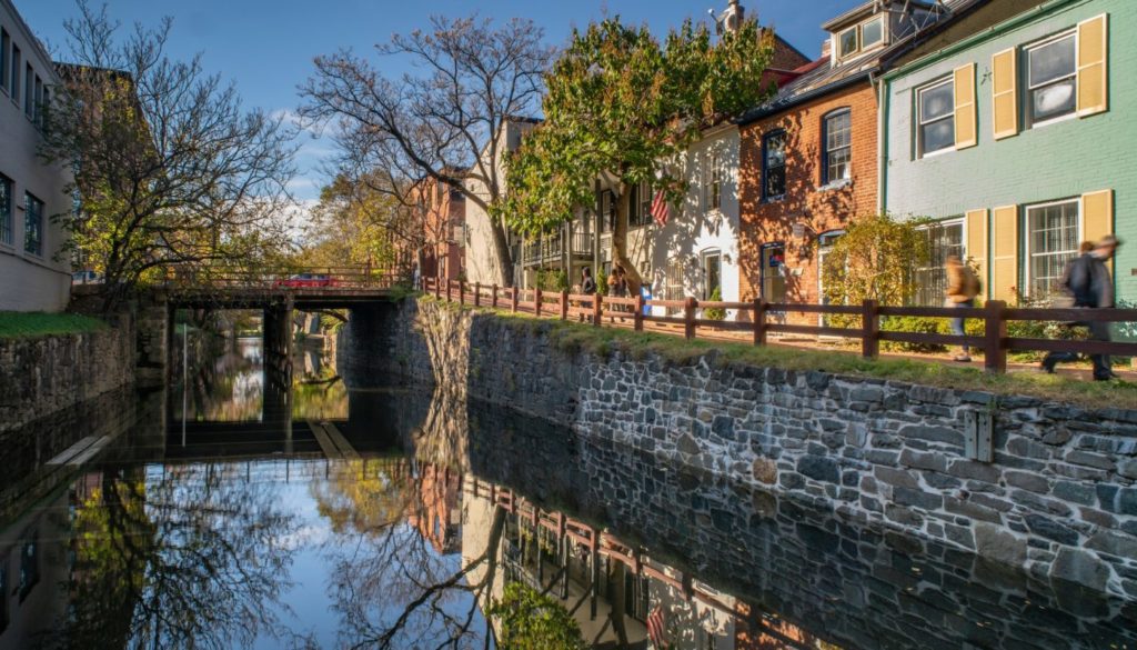 The C&O Canal Towpath in Georgetown, Washington, DC, with a walker passing historic rowhouses reflected in the still canal water