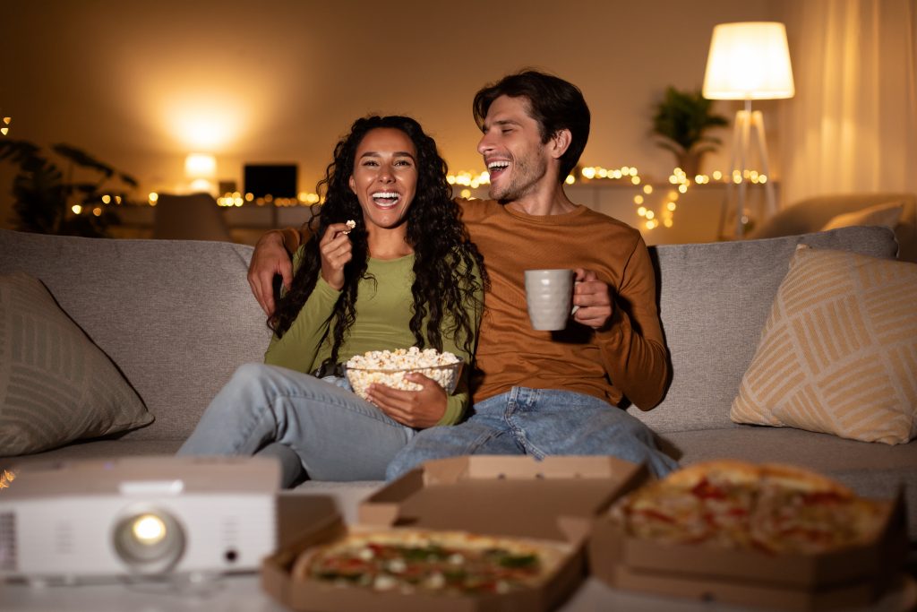 Stay In & Snuggle: A Heart-shaped Box of Romantic Movies Cheerful Couple Using Home Cinema Projector Watching Comedy Movie Indoors