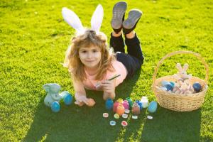 Child laying on grass in park wit easter eggs. Easter kids boy in bunny ears painting easter eggs outdoor. Cute child in rabbit costume with bunny ears having fun in park.