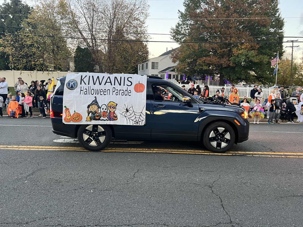 A car with a Kiwanis Halloween Parade banner drives past families in costume lining a Leesburg street