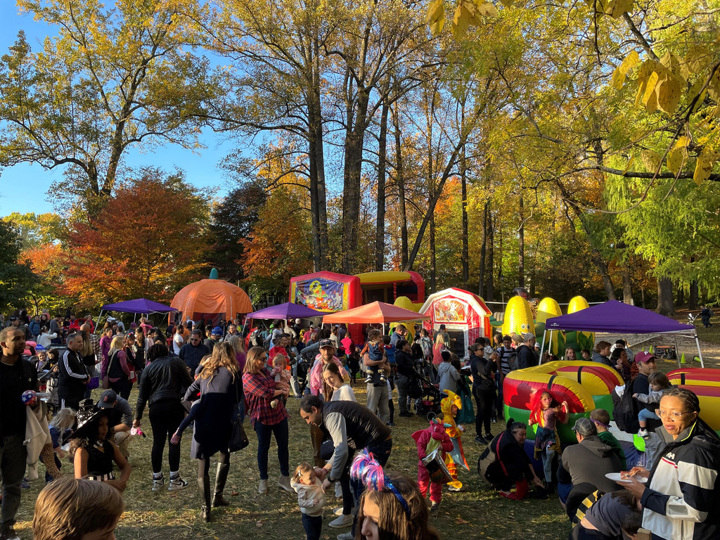 Families in costume crowd Cherry Hill Park under fall foliage at the Falls Church Halloween Carnival, with bounce houses in the background