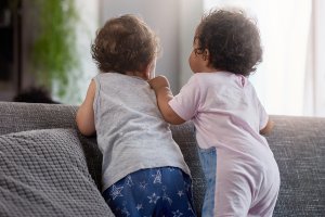 Back view of baby boy and girl standing together on back of couch, cute playing playdate interaction