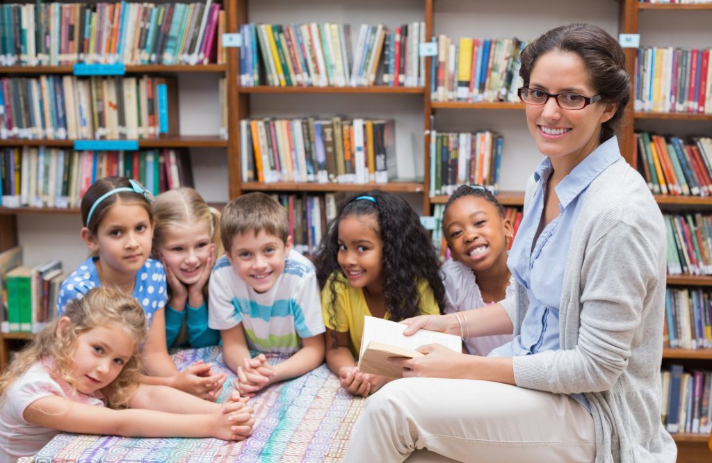 Honor your favorite teacher with these meaningful books for every age Cute pupils and teacher reading in library at the elementary school