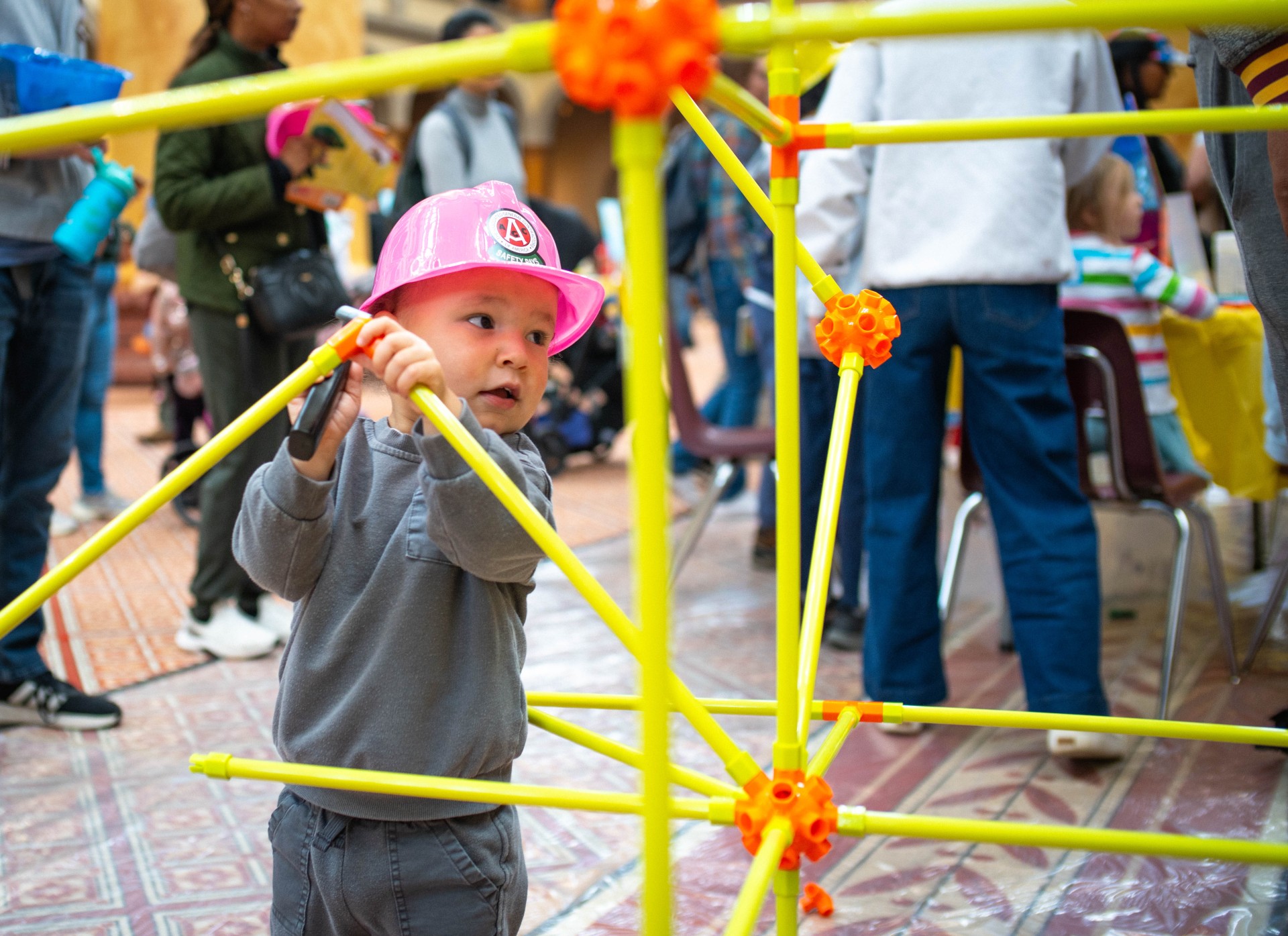 A toddler in a pink hard hat connects yellow poles into a large frame at The Big Build in Washington, DC.
