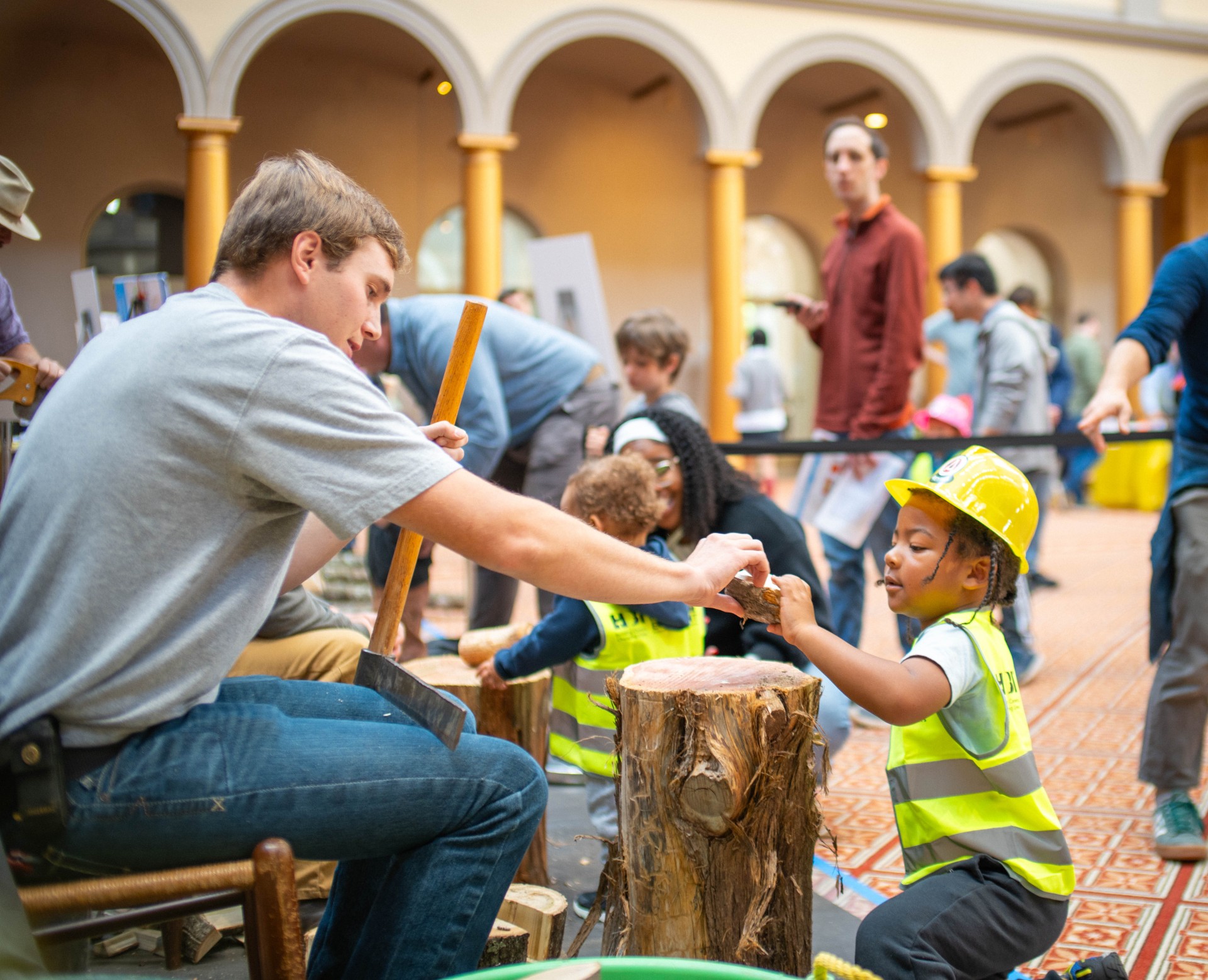 A volunteer hands a piece of split log to a young child in a hard hat and safety vest at a woodworking station.