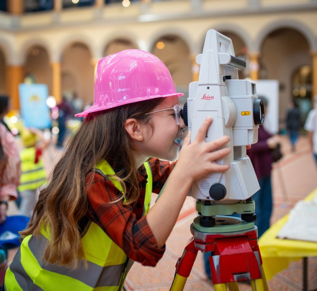 Step Into theWorld of Building at the National Building Museum’s The Big Build! A girl in a hard hat and safety glasses looks through a surveying instrument on a tripod at The Big Build.