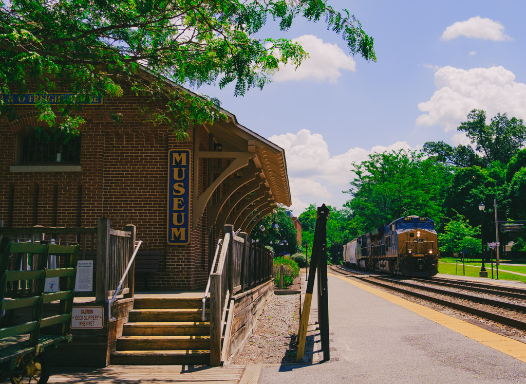 Brick Gaithersburg Community Museum building with a blue Museum sign beside the railroad tracks as a freight train approaches