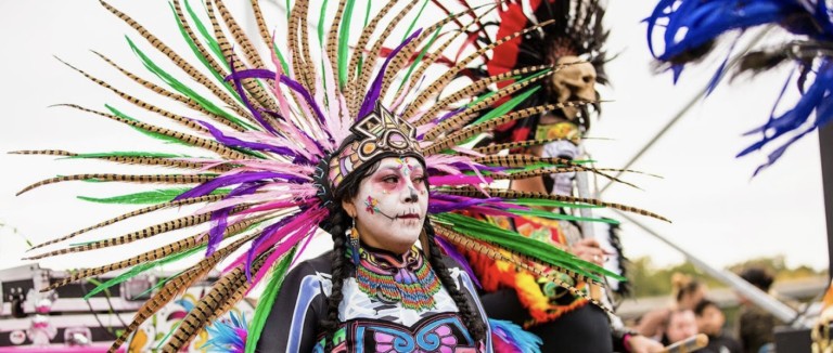 A dancer in a feathered headdress and sugar skull face paint performs at a Día de los Muertos festival