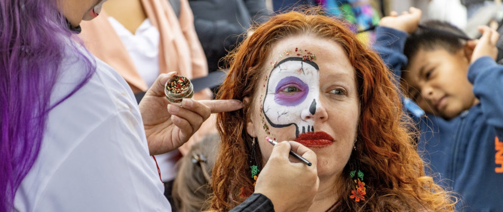 A face painter applies sugar skull makeup to a woman at a Día de los Muertos celebration
