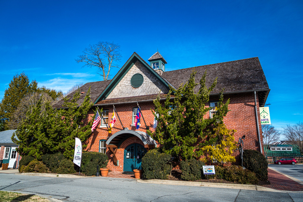 Red brick Arts Barn building in Gaithersburg with a green arched entrance, flags above the door and evergreen shrubs along the front