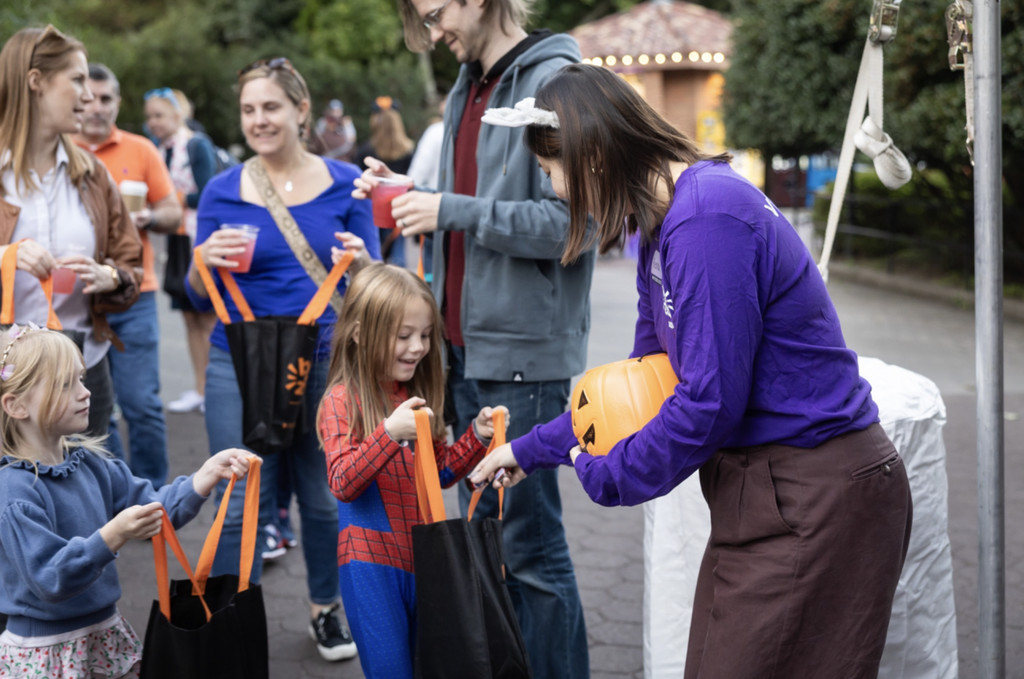 A zoo volunteer drops candy from a plastic pumpkin into a smiling girl’s treat bag at a Boo at the Zoo treat station.