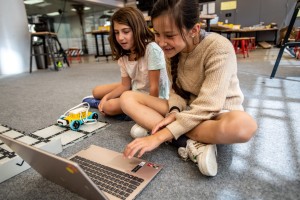 Two girls sit on a carpeted floor and use a laptop to program a small yellow and blue wheeled robot at KID Museum in Bethesda.