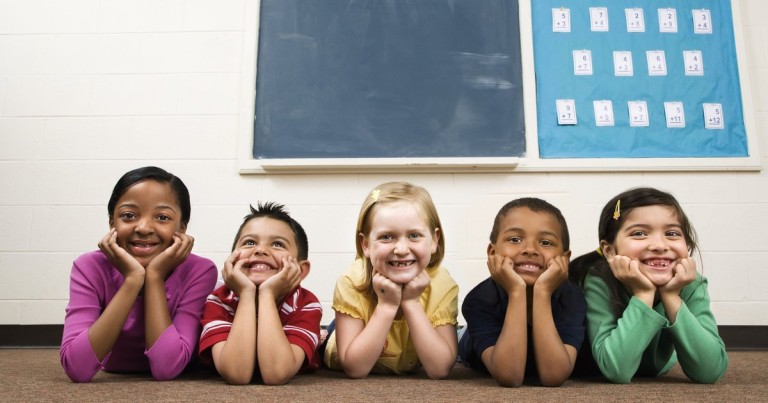 Five smiling elementary school kids lie on a classroom floor with their chins in their hands, ready for winter break camp.